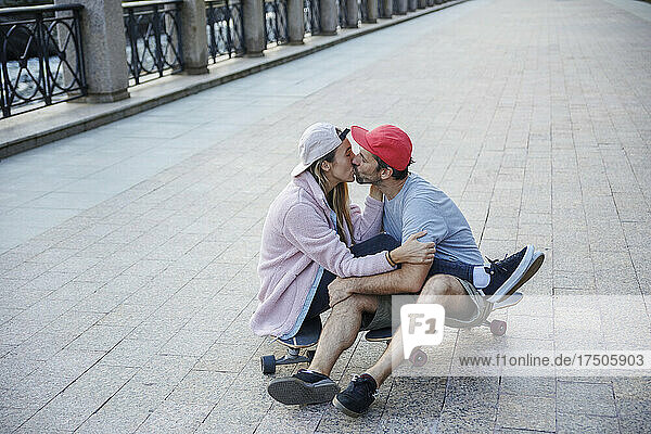 Affectionate couple kissing each other sitting on skateboard at footpath