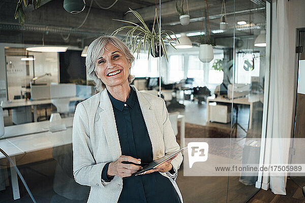 Senior businesswoman with tablet PC leaning on glass wall