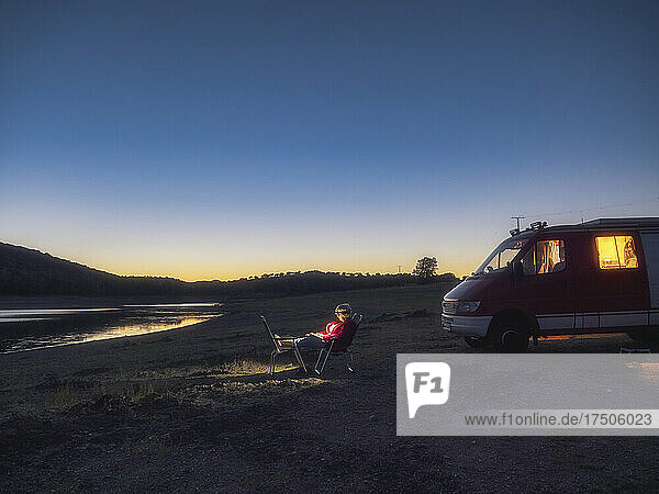 Woman sitting on chair near camper trailer at sunset
