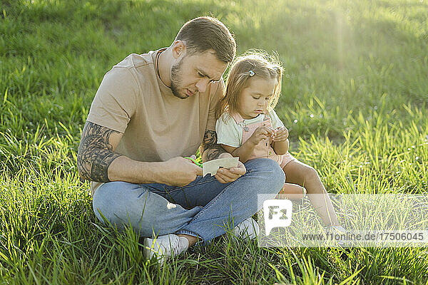 Mann  der Tätowierungen schneidet  sitzt im Schneidersitz seiner Tochter im Gras