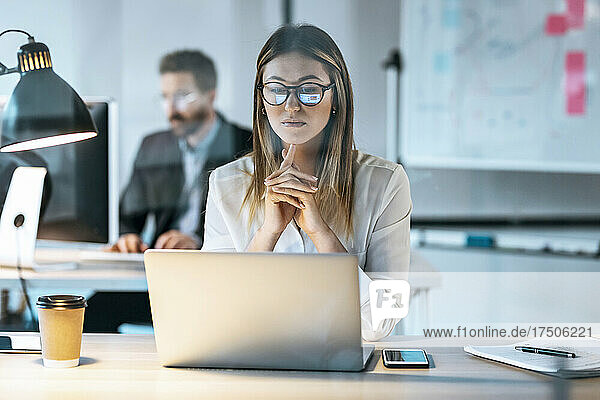 Nachdenkliche Geschäftsfrau blickt mit einem Kollegen im Hintergrund auf den Laptop