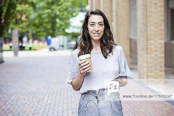 Lächelnde Geschäftsfrau hält Einwegbecher und Smartphone auf dem Fußweg
