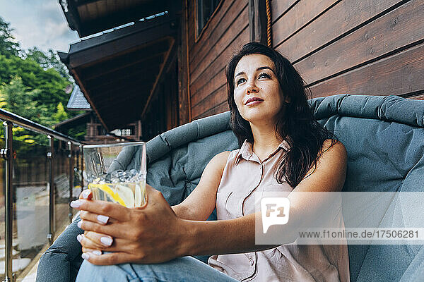 Nachdenkliche Frau mit Trinkglas sitzt auf einem Stuhl auf dem Balkon