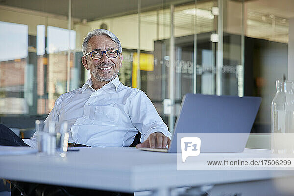 Happy mature businessman with laptop at table in office