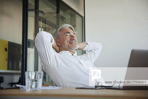 Smiling businessman relaxing with hands behind head at office