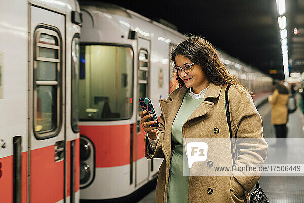 Smiling woman in overcoat using mobile phone at railroad station