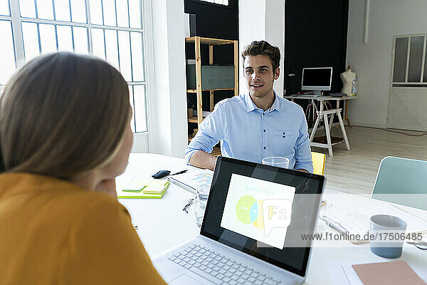 Young businessman talking to colleague with laptop at desk in office