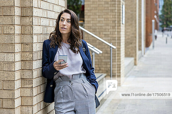 Businesswoman with smart phone leaning on brick wall