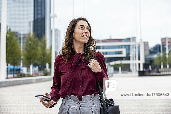 Contemplating businesswoman holding smart phone walking on footpath in city