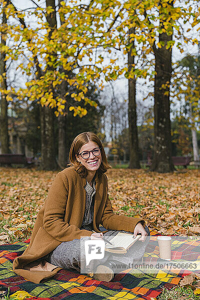 Smiling woman with book sitting on blanket in autumn park