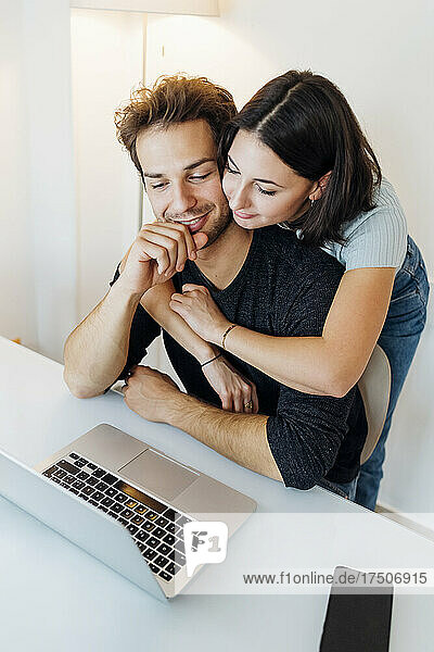 Junges Paar umarmt sich  während es zu Hause am Laptop arbeitet