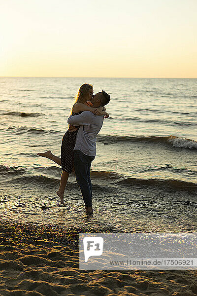 Romantischer Mann hebt und küsst Frau am Strand bei Sonnenuntergang