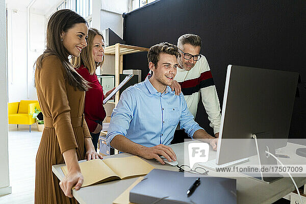 Young businessman using desktop PC with colleagues standing at desk in office