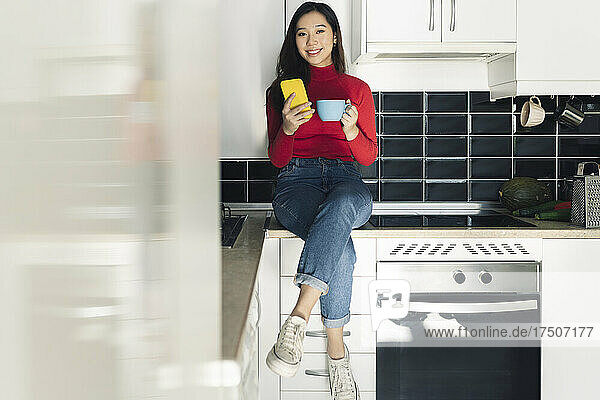 Smiling woman with coffee cup and mobile phone at kitchen counter