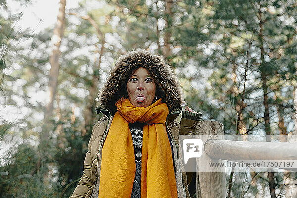 Woman in fur cap sticking out tongue by railing at forest