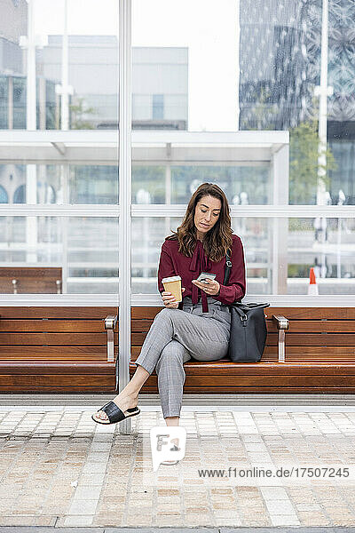 Businesswoman with disposable cup using smart phone waiting at railroad platform
