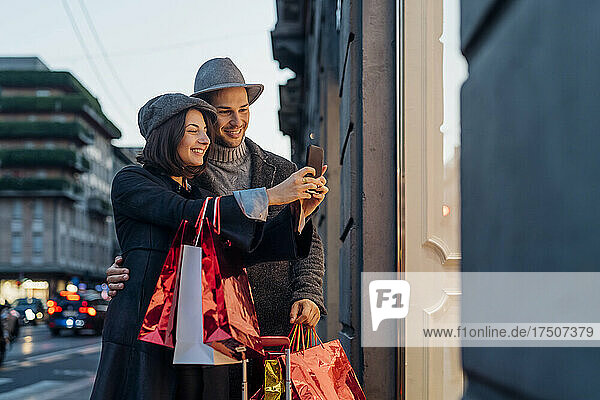 Lächelndes junges Paar mit Einkaufstüten  das in der Stadt ein Selfie mit dem Smartphone macht