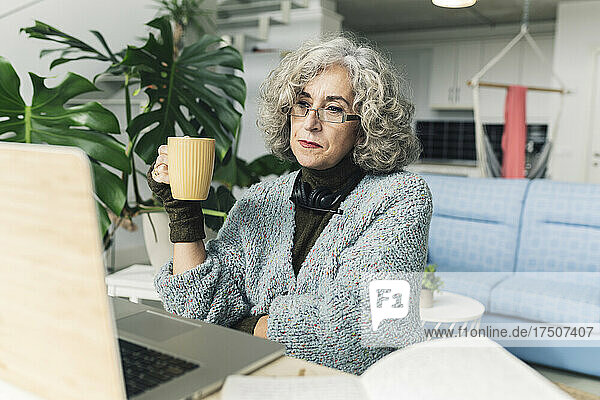 Ältere Geschäftsfrau hält Kaffeetasse vor Laptop im Heimbüro