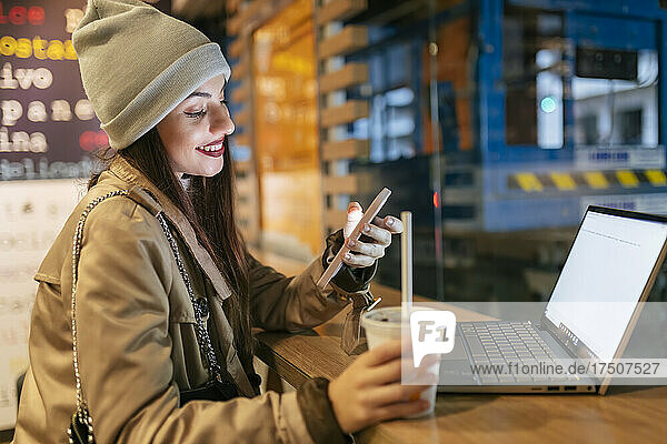 Woman using smart phone holding drink by laptop at cafe