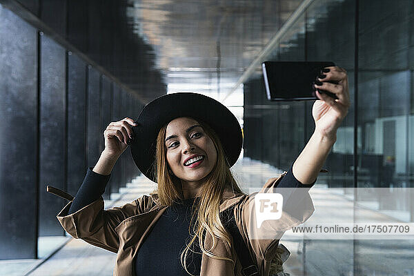 Smiling woman with hat taking selfie through mobile phone on footpath