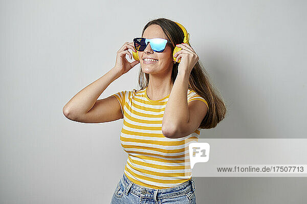 Woman with sunglasses listening music through headphones against gray background