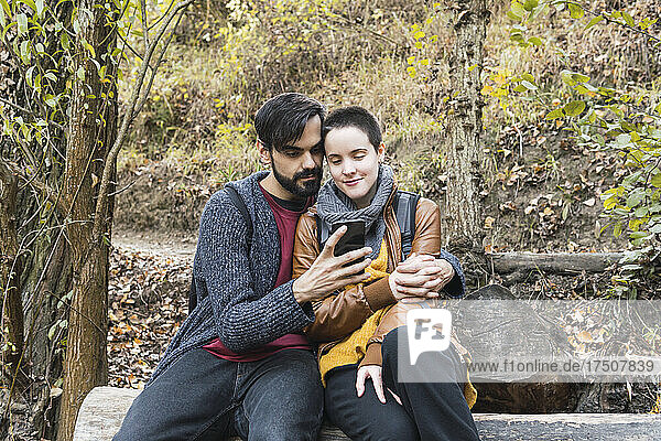 Bearded man sharing smart phone with girlfriend on log at riverbank