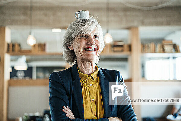 Happy businesswoman with arms crossed and coffee cup on head in office