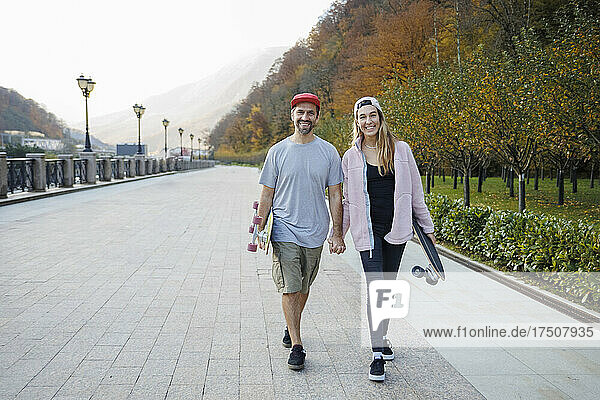Lächelndes Paar mit Skateboards  die Händchen haltend gemeinsam auf dem Fußweg spazieren gehen