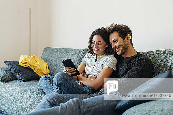 Smiling girlfriend sharing smart phone with boyfriend on sofa at home