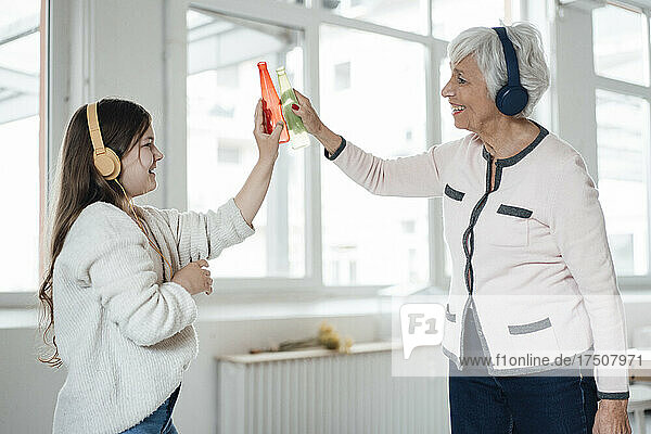 Woman and granddaughter with headphones toasting bottles at home