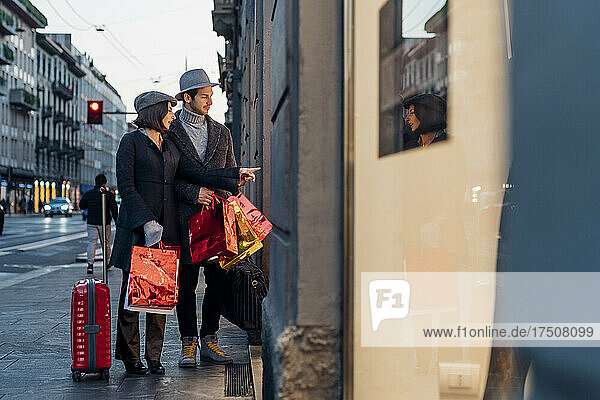 Young couple with wheeled luggage window shopping in city