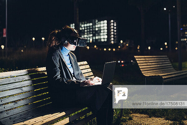 Businesswoman with virtual reality headset using laptop on bench at night
