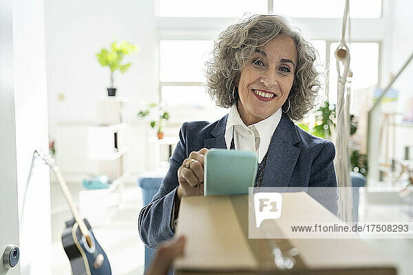 Senior businesswoman with mobile phone receiving package at home