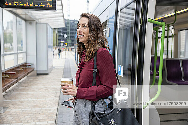 Lächelnde Geschäftsfrau mit Einwegbecher und Mobiltelefon steigt am Bahnhof aus dem Zug