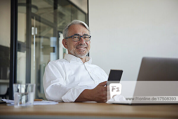 Smiling mature businessman with smart phone and laptop in office