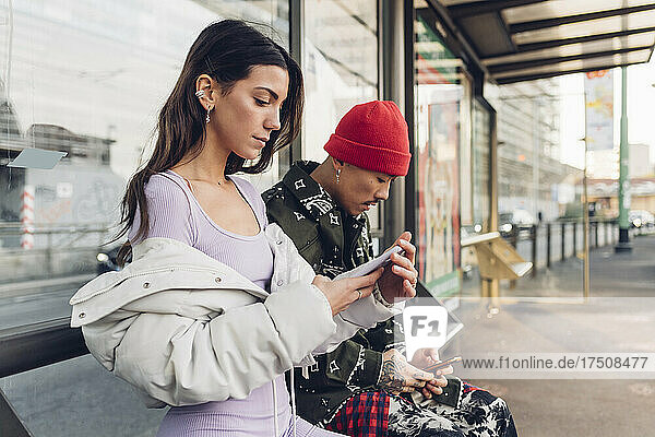 Paar sitzt mit Smartphones auf Bank an Bushaltestelle