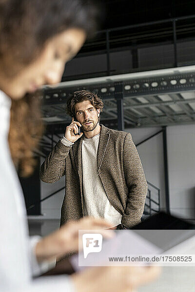 Businessman talking on mobile phone with colleague working in foreground at industrial hall