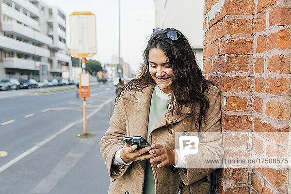 Smiling curvy woman using mobile phone while leaning on brick wall