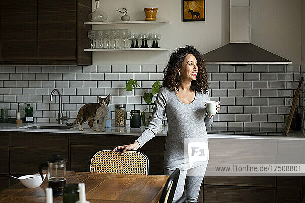 Lächelnde Frau mit Kaffeetasse  die zu Hause in der Küche wegschaut