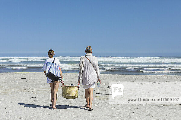 Mutter und Tochter im Teenageralter gehen am Sandstrand spazieren und tragen einen Korb
