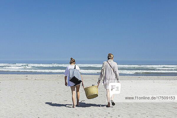 Mother and teenage daughter walking on a sandy beach carrying a basket