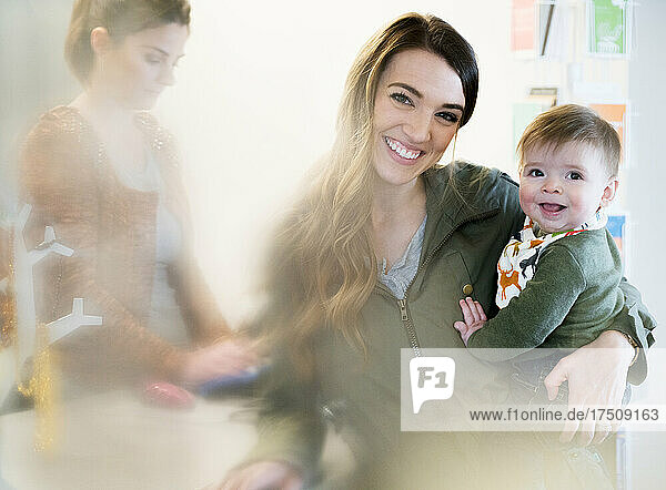 Frau mit Baby neben einer Registrierkasse mit Verkäuferin  die in die Kamera lächelt.
