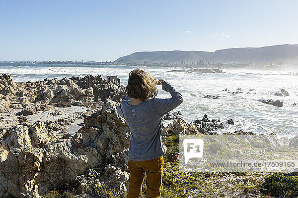 A boy on the rocky shore looking over the surf and sea spray.