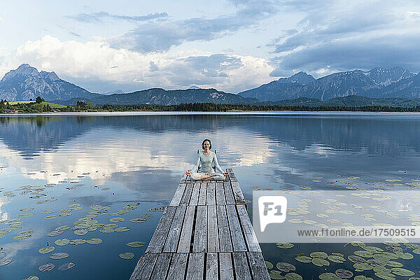 Woman meditating while sitting on jetty over lake against cloudy sky