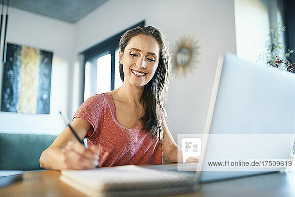 Smiling female freelancer writing in note pad on desk at home office