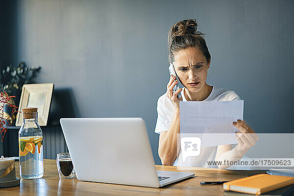 Businesswoman holding document talking over smart phone while sitting at desk in home office