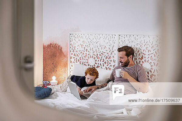 Boy looking at laptop being used by father on bed