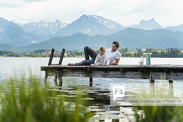 Father with daughter relaxing on jetty over lake against mountains