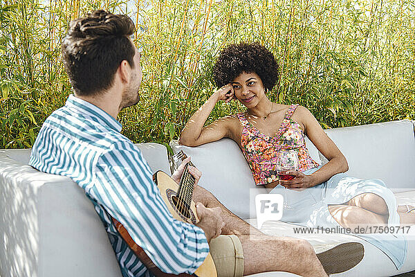 Woman holding red wine glass while looking at man playing guitar on sofa at penthouse patio