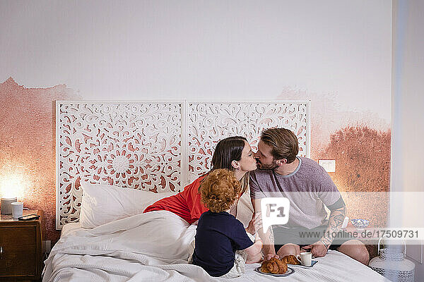 Parents kissing while having breakfast with son bed at home